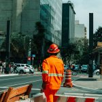 Trabalhador da construção civil em uma rua movimentada de São Paulo, cercado por edifícios e circulação urbana.