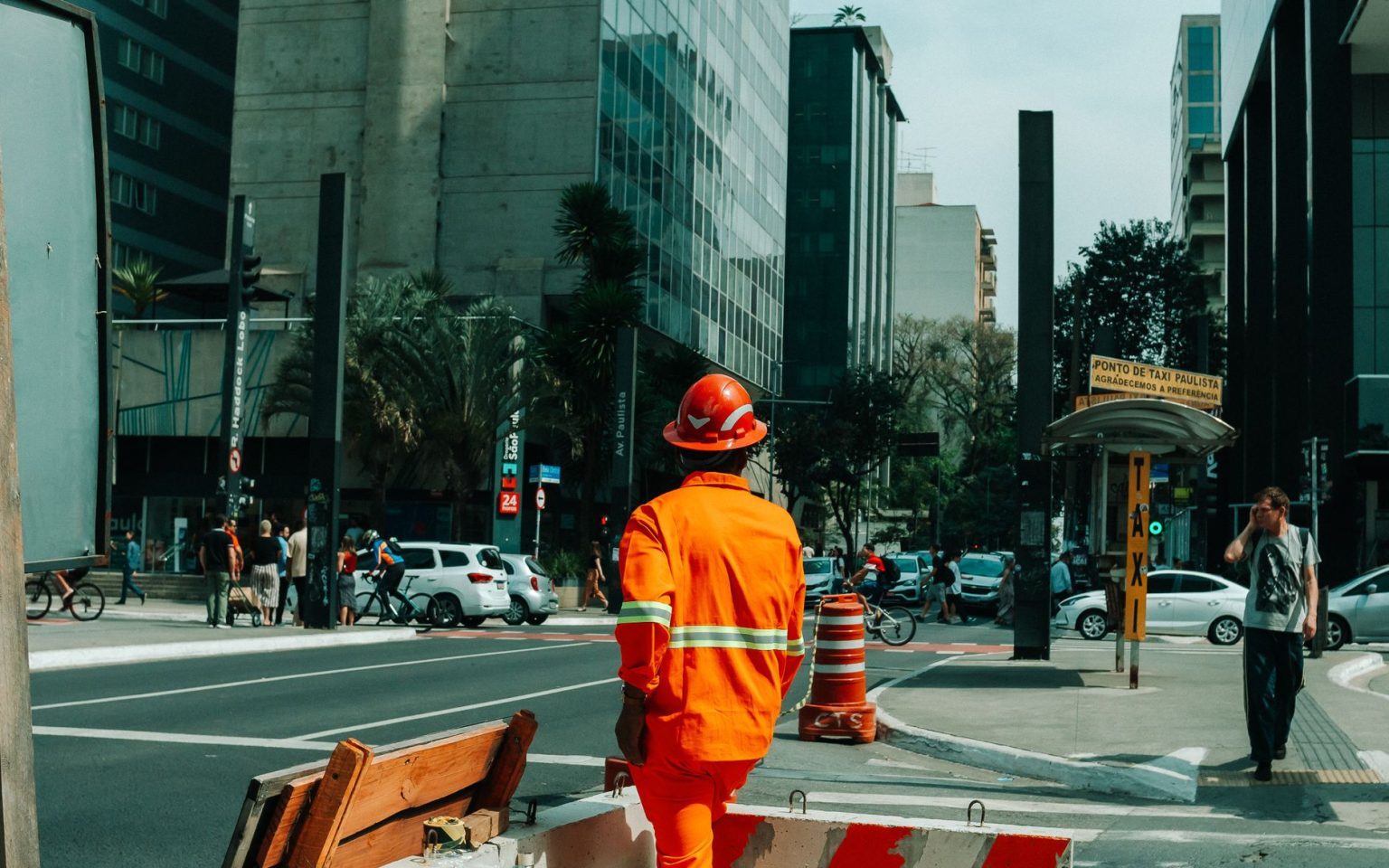 Trabalhador da construção civil em uma rua movimentada de São Paulo, cercado por edifícios e circulação urbana.