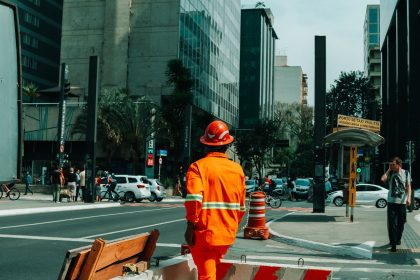 Trabalhador da construção civil em uma rua movimentada de São Paulo, cercado por edifícios e circulação urbana.