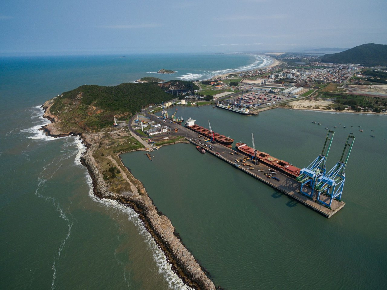 Vista aérea do Porto de Imbituba, em Santa Catarina, com docas, guindastes, navios e contêineres.