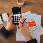 Mãos usando um aplicativo de calculadora no celular sobre documentos financeiros.