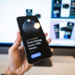 Mão segurando um smartphone com a tela de um aplicativo de carteira digital, com um monitor desfocado ao fundo