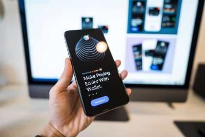 Mão segurando um smartphone com a tela de um aplicativo de carteira digital, com um monitor desfocado ao fundo